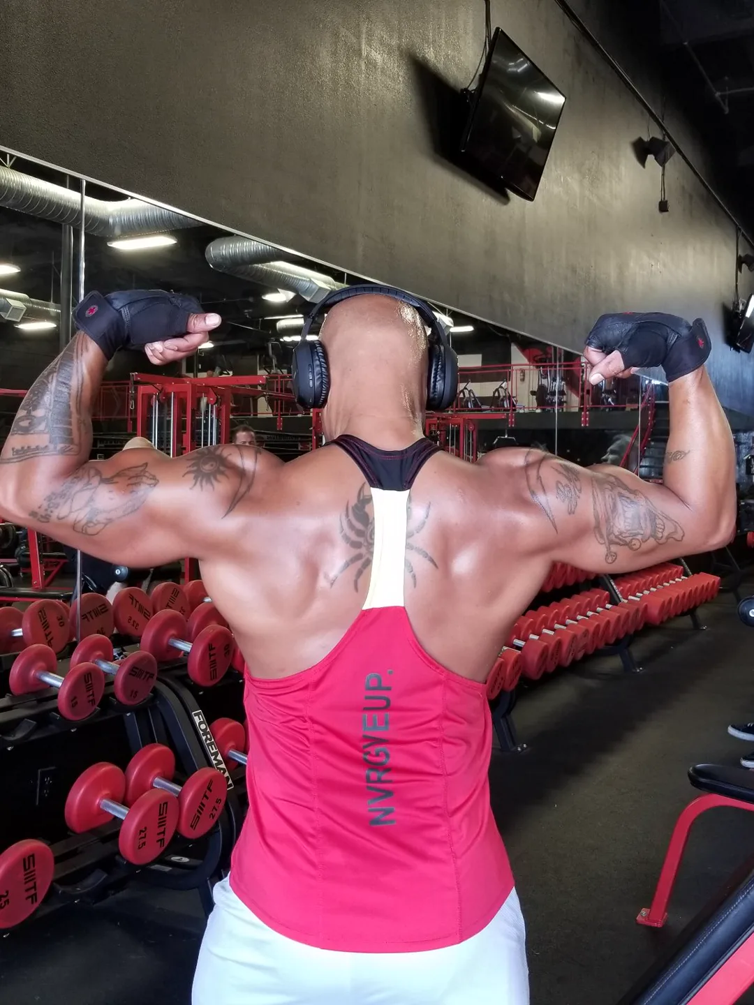 Coach Bizz hitting a back double-biceps flex in front of the dumbbell rack